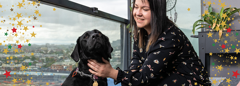 Brittee is smiling and patting her black Labrador Seeing Eye Dog, on a balcony with urban views